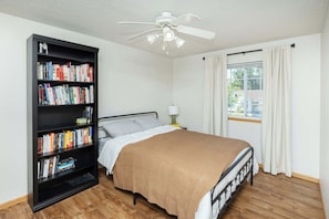 Ein Schlafzimmer mit Bett, Bücherregal, Deckenventilator, einem Fenster mit Vorhängen und einem Nachttisch mit Lampe.