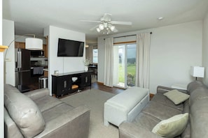 Un salon moderne avec un canapé, un téléviseur, un ventilateur de plafond et, au fond, un coin cuisine.