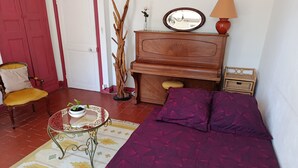 A cozy room with a wooden piano, a sofa with a purple cover, a small table with a plant, and a chair with a yellow cushion. - Le soleil rayonnant: bed and breakfast at the foot of Vézelay hill, (Asquins)