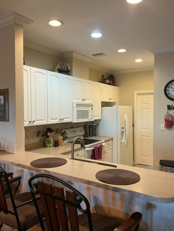 A kitchen with white cabinets, a microwave, stove, and a refrigerator. There are two bar stools and a clock on the wall. - Sunsets, Canal Front, FISH, FISH, FISH! Suwannee Cove - Unit 207, Boat Slip 23 (Suwannee)