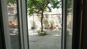 A view through a window of a courtyard with a tree, potted plants, and a stone wall. - Casa Ammare - One Bedroom House, Sleeps 2 (Syracuse)