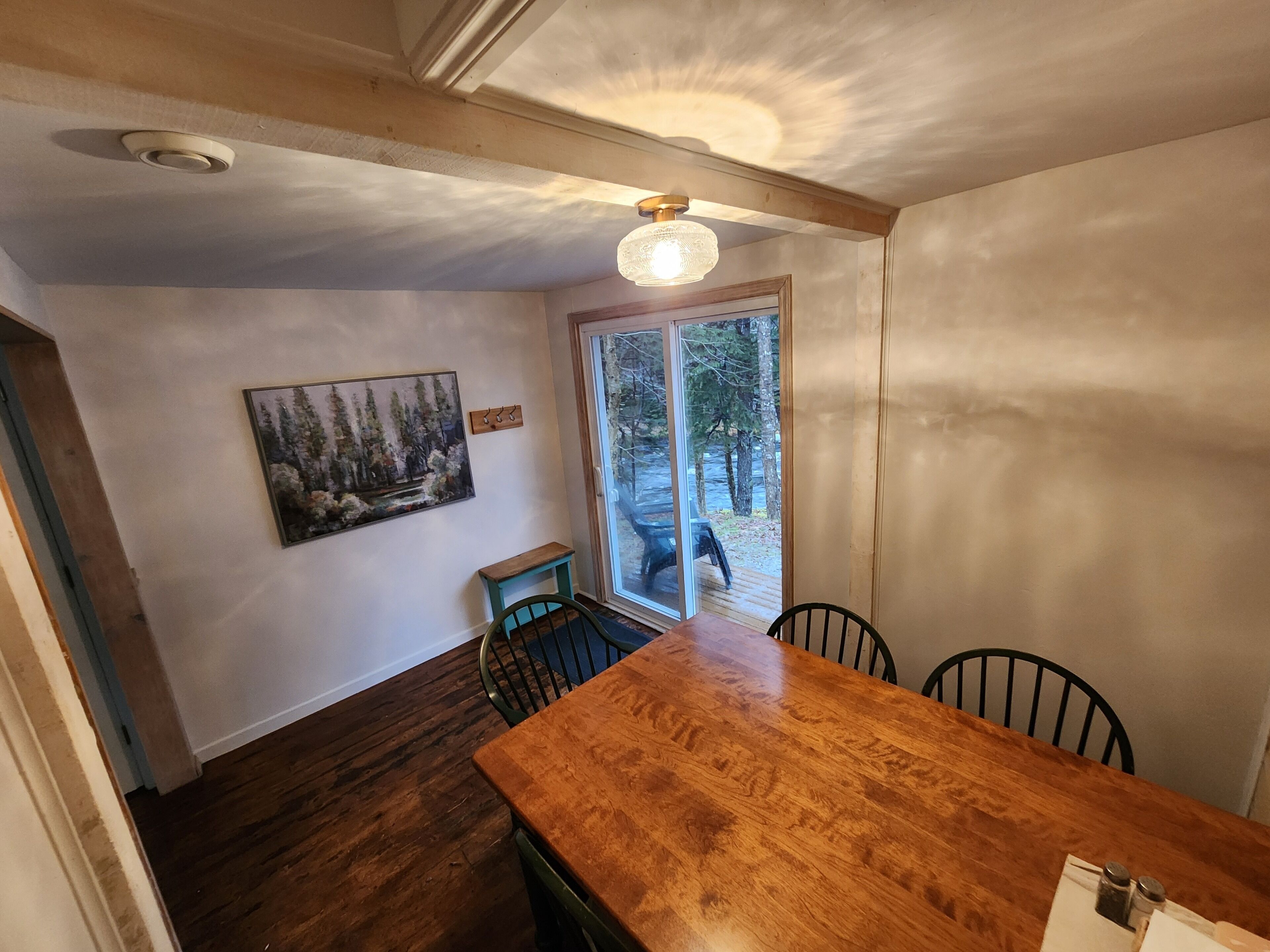Un comedor con una mesa y sillas de madera, un ventilador de techo, un cuadro de un bosque y un divisorio de vidrio corredizo que da a un espacio exterior.