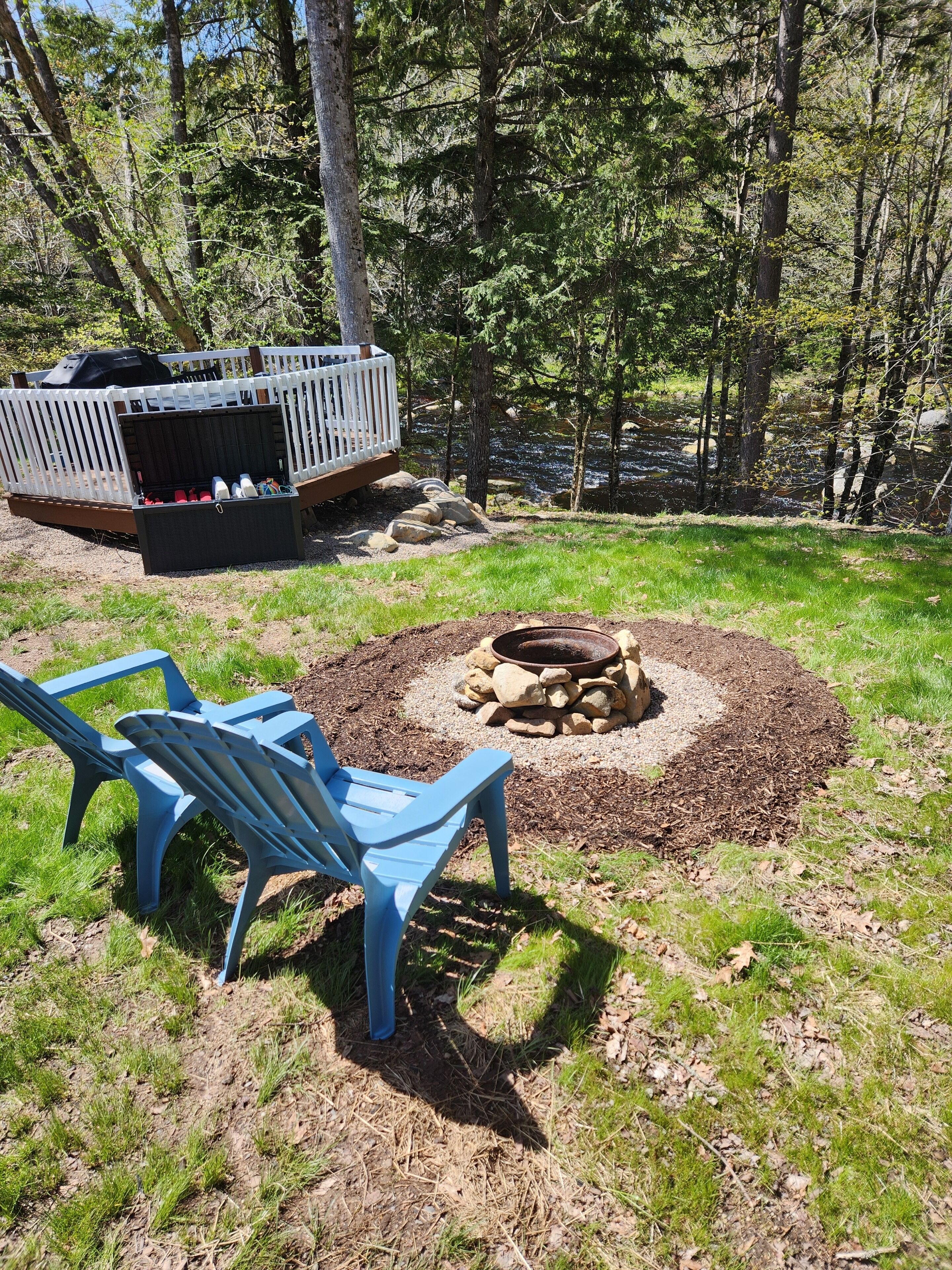 A backyard with a fire pit, two blue chairs, and a white deck.