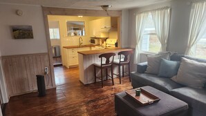 A cozy living room with a grey sofa and wooden flooring. A kitchen with a wooden island and bar stools is visible in the background.
