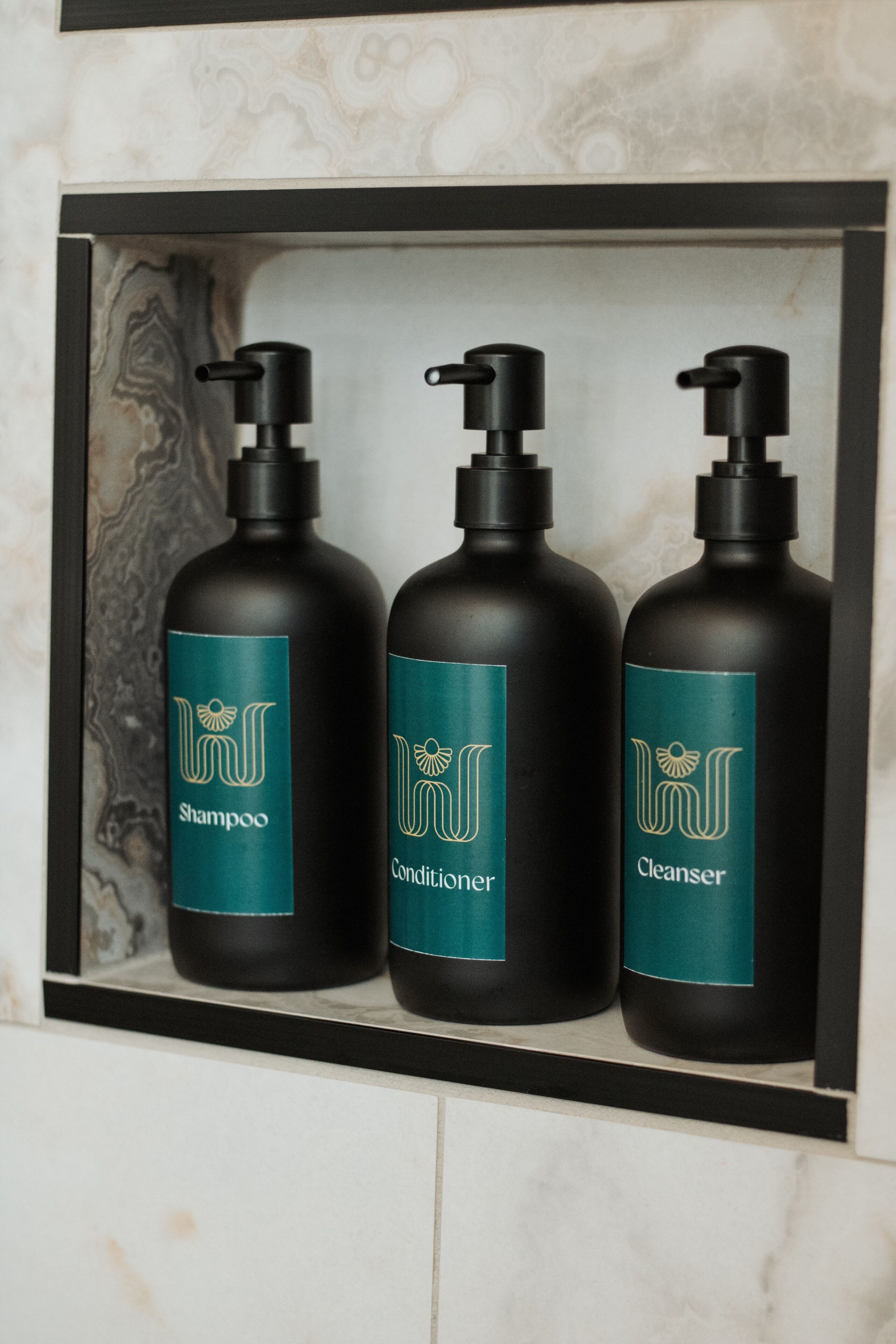 Uma prateleira no banheiro com três frascos pretos rotulados como shampoo, condicionador e limpador.