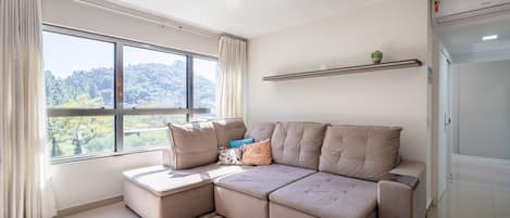 Una sala moderna con un sofá seccional, un ventanal con cortinas, un tapete estampado y una planta pequeña en un estante.