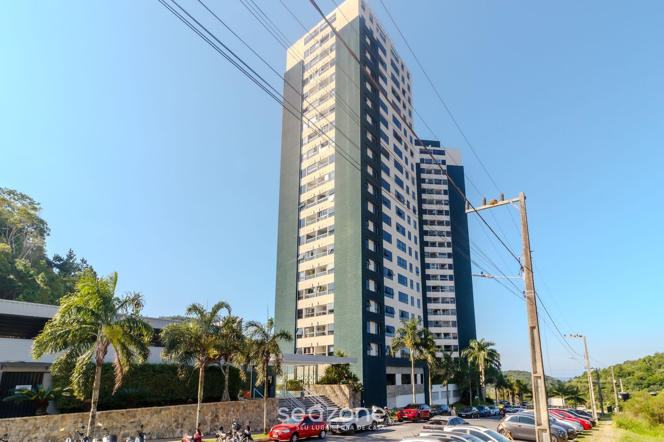 A tall residential building with multiple balconies, surrounded by parked cars and palm trees.