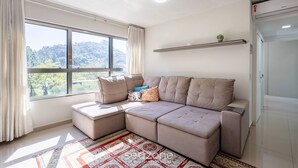 A modern living room with a sectional sofa, a large window with curtains, a patterned rug, and a small plant on a shelf. - Beautiful Apartment in Building With Pool Mpb0034 (Itajai)