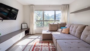 A modern living room with a large window, a flat-screen TV, a sofa, and a patterned rug. - Beautiful Apartment in Building With Pool Mpb0034 (Itajai)
