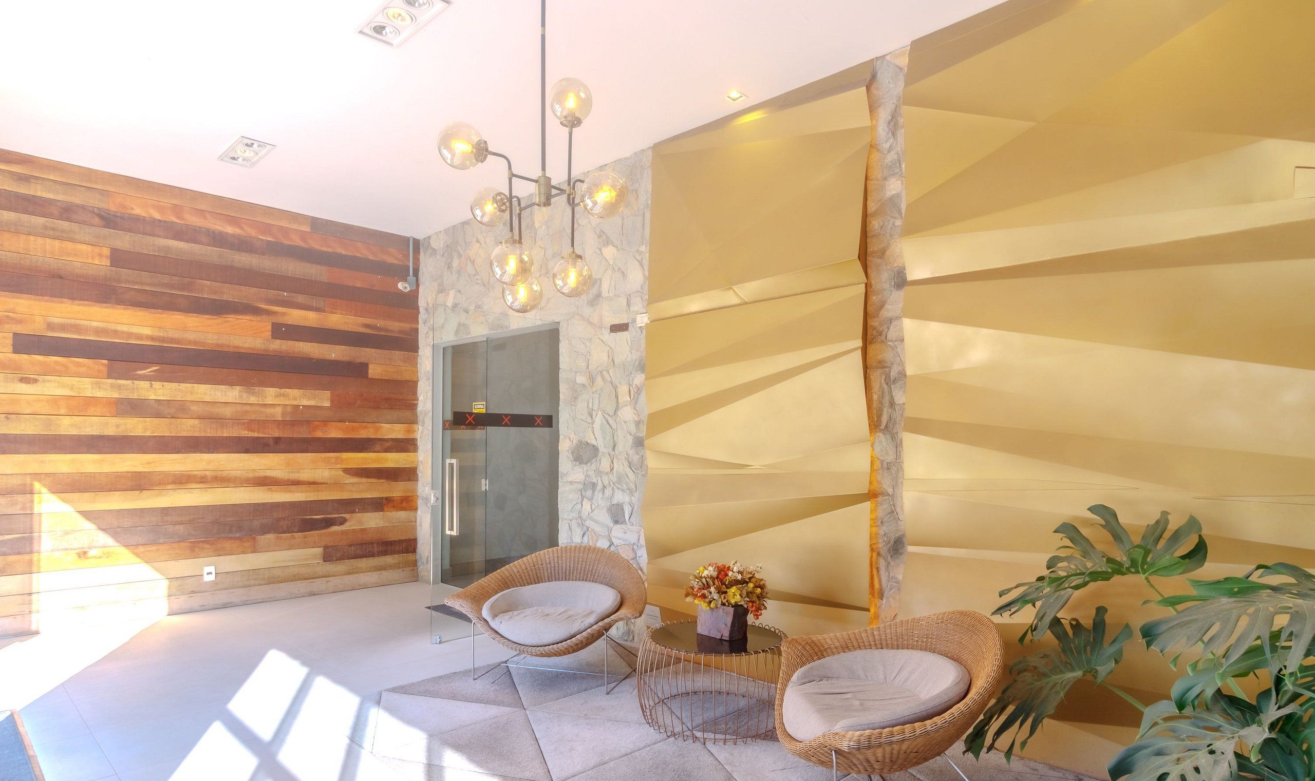 Un interior moderno con paneles de madera, una pared de acento en piedra y una puerta de vidrio que da al baño. Hay dos sillas de mimbre y una mesita con un florero.