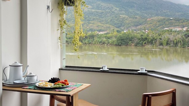 Une pièce avec vue sur un lac et des montagnes, une table avec une théière et des tasses, deux chaises en bois et une plante suspendue.
