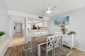 A modern kitchen with white cabinets, a dining area with a wooden table and white chairs, and a ceiling fan. - Oceanfront 2BR Condo Bright Breezy Beachfront Indian Harbour Beach, 606 (Indian Harbour Beach)