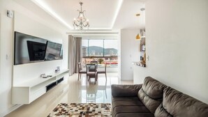 A modern living room with a flat-screen TV, a brown leather sofa, and a dining area with a view of buildings and mountains. - 2BR Apt With BBQ in Meia Praia Mlo0601 (Itapema)