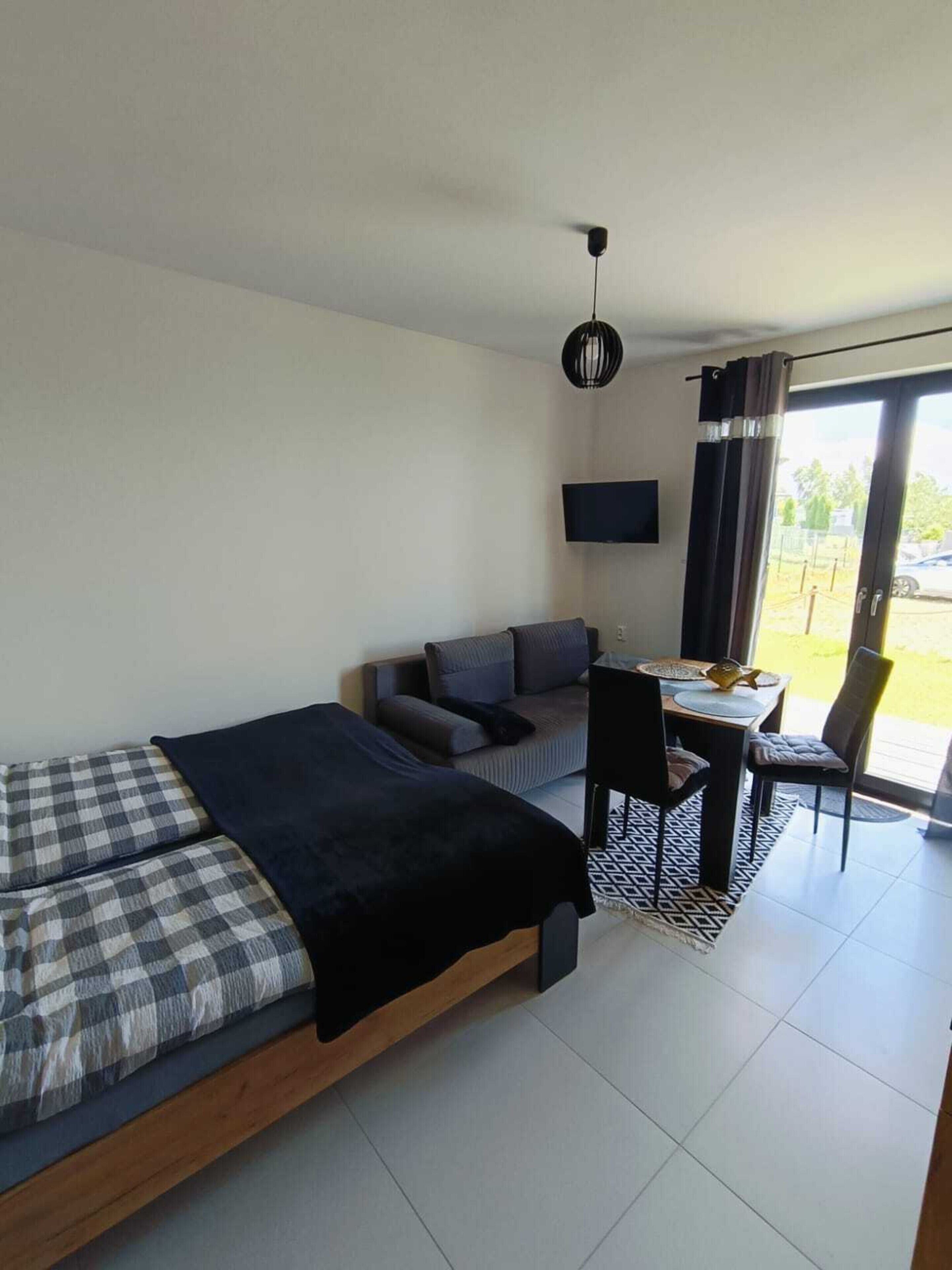 A modern bedroom with a bed, a sofa, a dining table with chairs, a TV, and a large window with curtains.