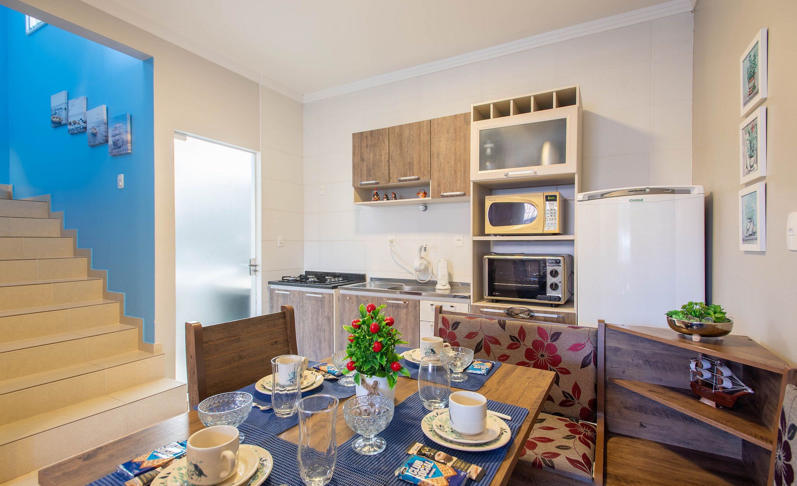 Una cocina moderna con armarios de madera, un microondas y un refrigerador. Hay una mesa de comedor con platos, tazas y un jarrón de flores.