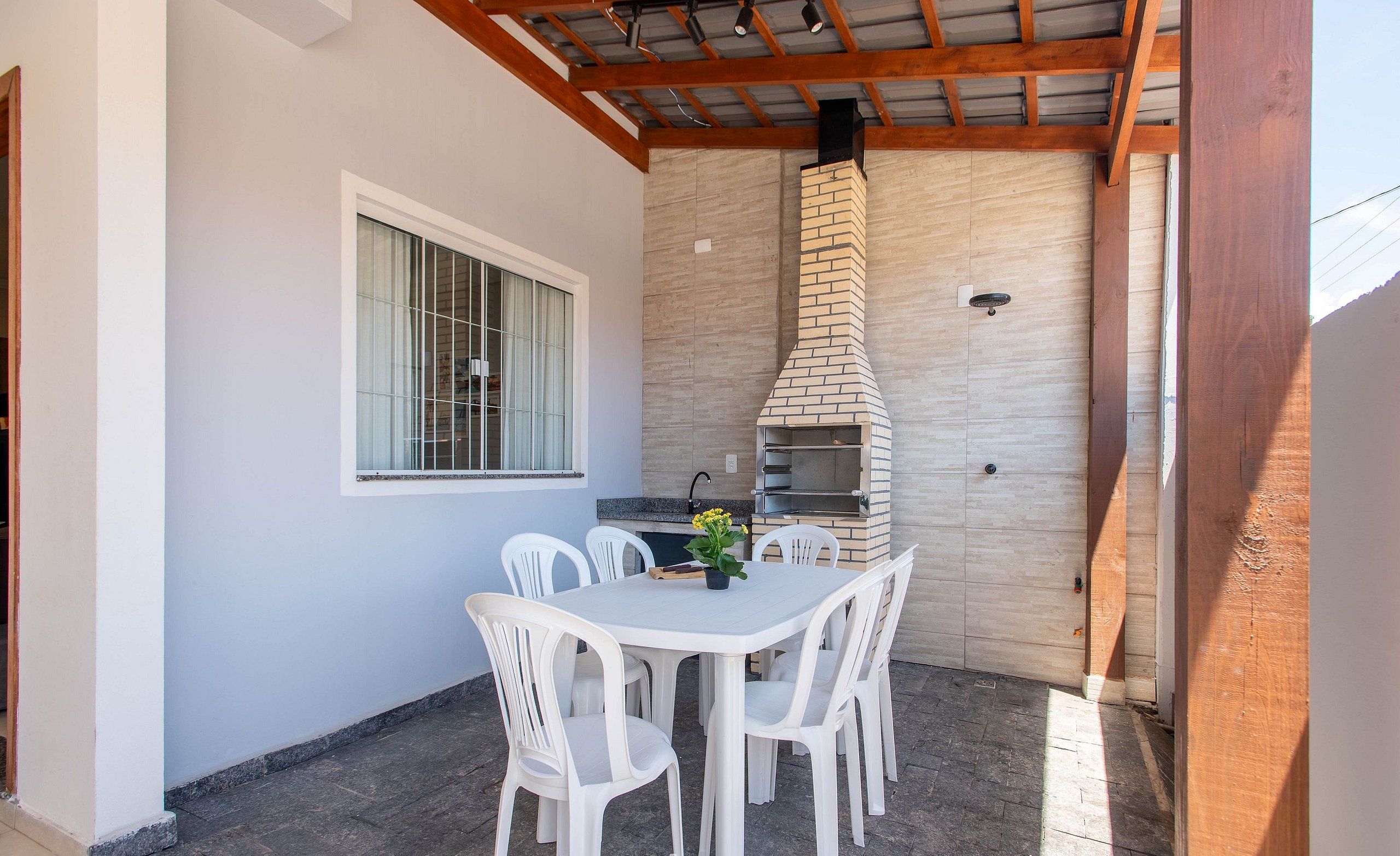 Une terrasse extérieure avec une table et des chaises blanches, une cheminée en briques et une fenêtre avec des stores.