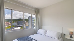 A hotel room with a bed, a bedside table with a lamp, and a window offering a city view. - Modern Apartment Near the BC Ferris Wheel Mbh0601 (Balneário Camboriú)