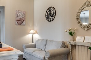 A room with a sofa, a wall clock, a mirror, a lamp, and a vase with flowers.