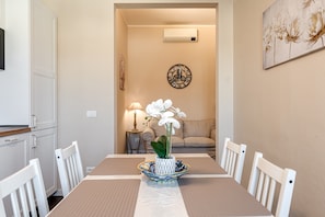 A dining area with a table set for four, a vase with flowers, a clock on the wall, and a sofa in the background.