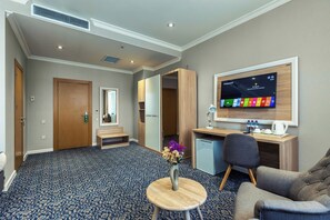 A hotel room with a large flat-screen TV mounted on the wall, a desk with a chair, a small refrigerator, a bench, and a patterned carpet. - METRO CITY HOTEL (Baku)