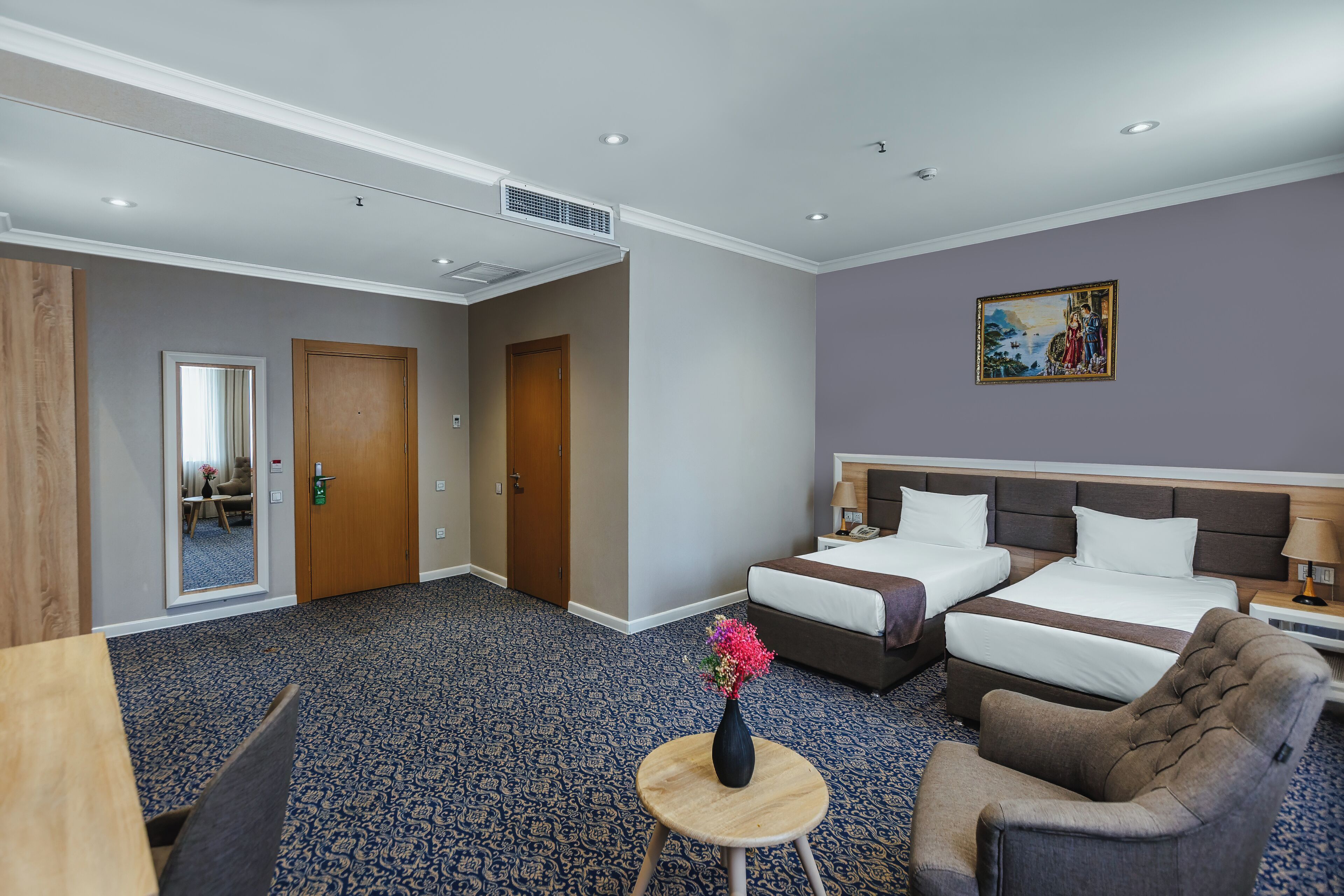 Ein Hotelzimmer mit zwei Betten, einer Sitzecke mit einem Sessel, einem kleinen Tisch mit einer Vase und einem Bild an der Wand.