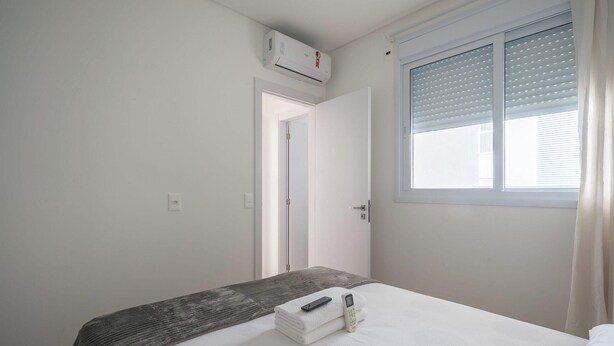 A bedroom with a bed, a bedside table, a window with blinds, an air conditioning unit, and a door.
