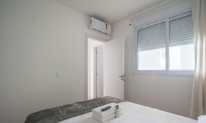 Une chambre d’hôtel comprenant un lit, une table de chevet avec une télécommande et un téléphone, une fenêtre avec des stores et un appareil de climatisation.
