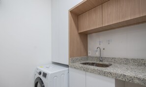 Een wasruimte met een wasmachine, een granieten aanrechtblad, een wasbak en houten kastjes.