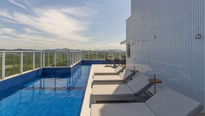 Una piscina infinita en la azotea con sillas de descanso y una mesa, rodeada por un edificio blanco con vistas al paisaje.