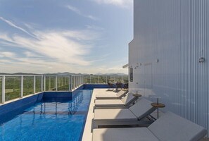 Une piscine à débordement située sur le toit, avec des chaises longues et une table, le tout entouré d’un bâtiment blanc offrant une vue sur le paysage.