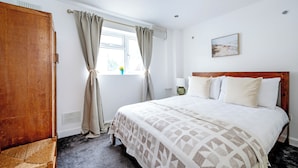 A bedroom with a wooden headboard, a large bed with a patterned bedspread, a window with curtains, a bedside lamp, and a wall-mounted picture. - 2 Bedroom flat in Putney by the river (London)