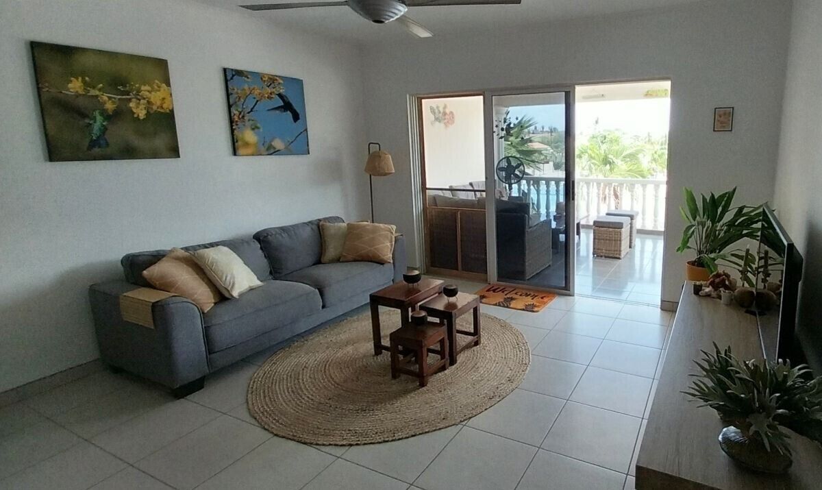 A living room with a grey sofa, two paintings, a small wooden table, and a large window with a view of a balcony.