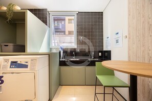 A compact kitchen with a washing machine, a sink, a window, and a green chair. - Charming studio in São Caetano ap111 (São Caetano do Sul)
