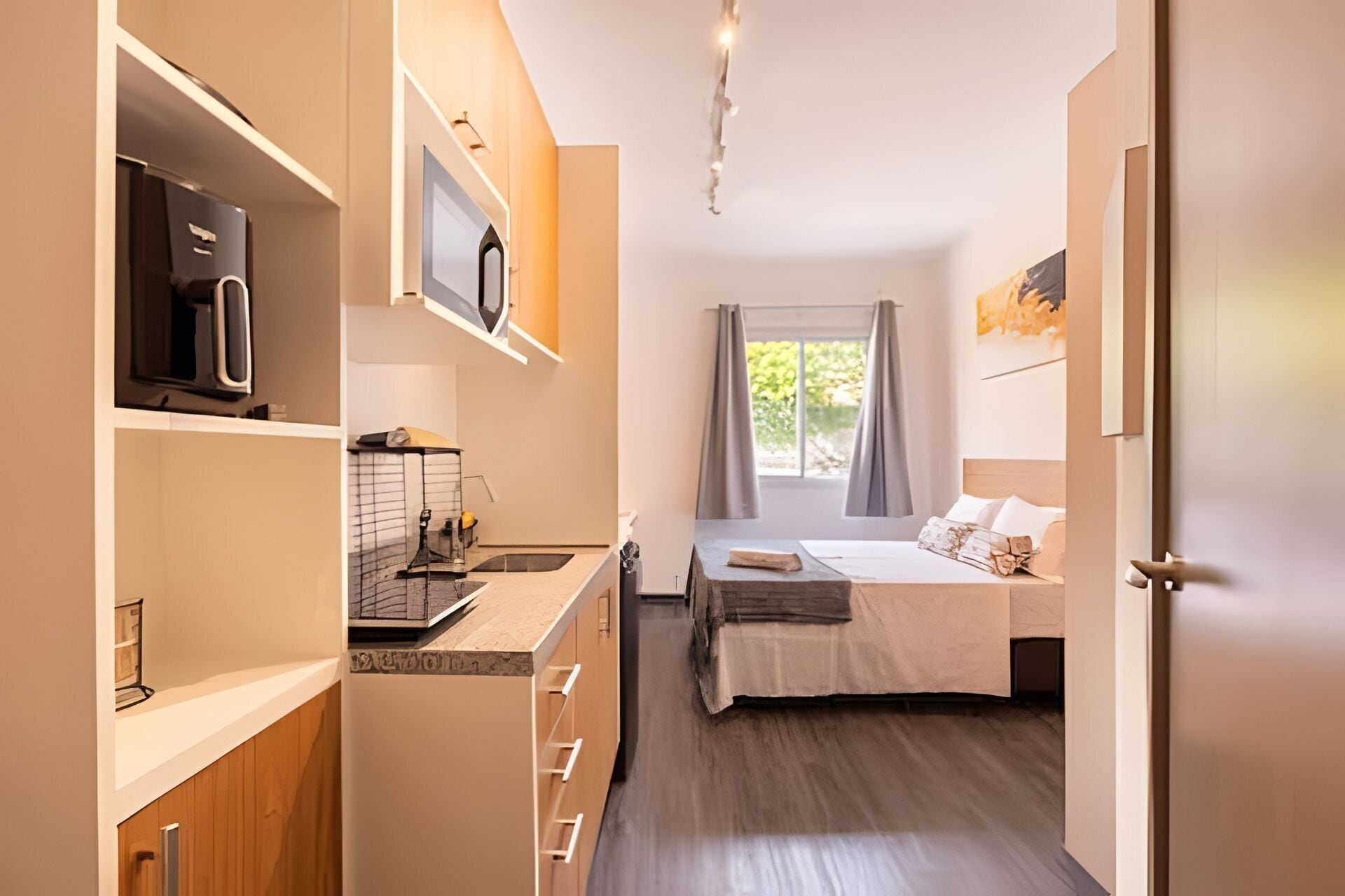A compact hotel room with a kitchenette, a bed with a wooden headboard, and a window with curtains.