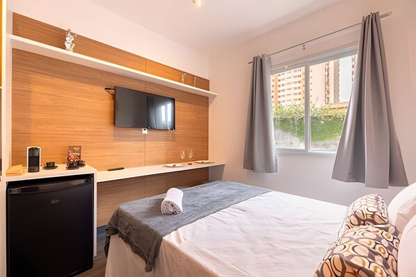 A hotel room with a bed, a desk with a flat-screen TV, a small refrigerator, and a window with curtains.