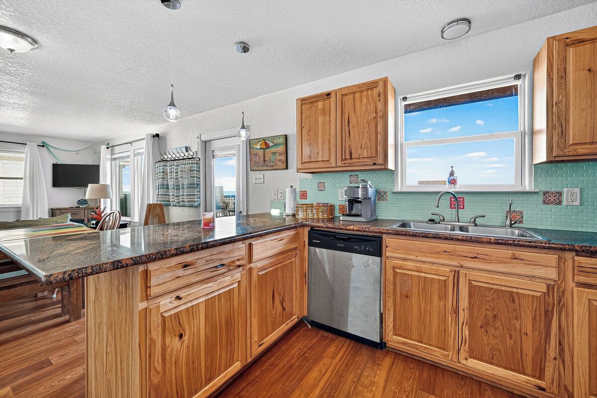 Una cocina con armarios de madera, lavavajillas, ventana con vistas y encimera con diversos objetos.