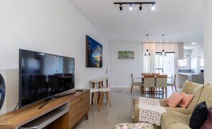 A modern living room with a flat-screen TV, wooden furniture, a dining area with chairs, and a kitchen in the background. - Cozy Apt 550m From the Beach in Porto Belo Lnb502 (Porto Belo)