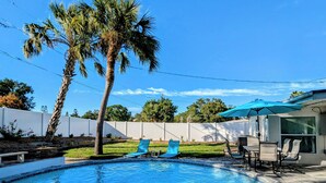 A swimming pool with two lounge chairs and a patio table, surrounded by a white fence and palm trees.