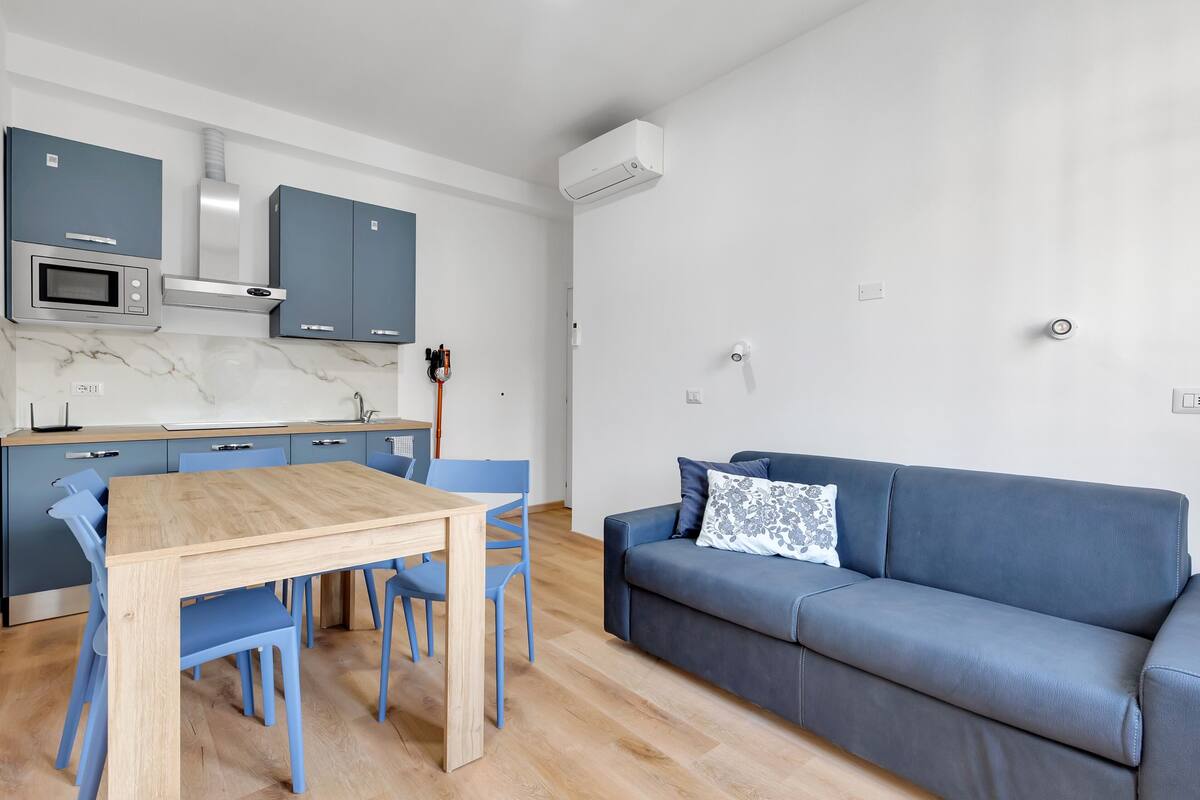 Uma cozinha moderna com uma mesa de madeira e cadeiras azuis, um sofá azul com almofada estampada e um aparelho de ar condicionado branco na parede.