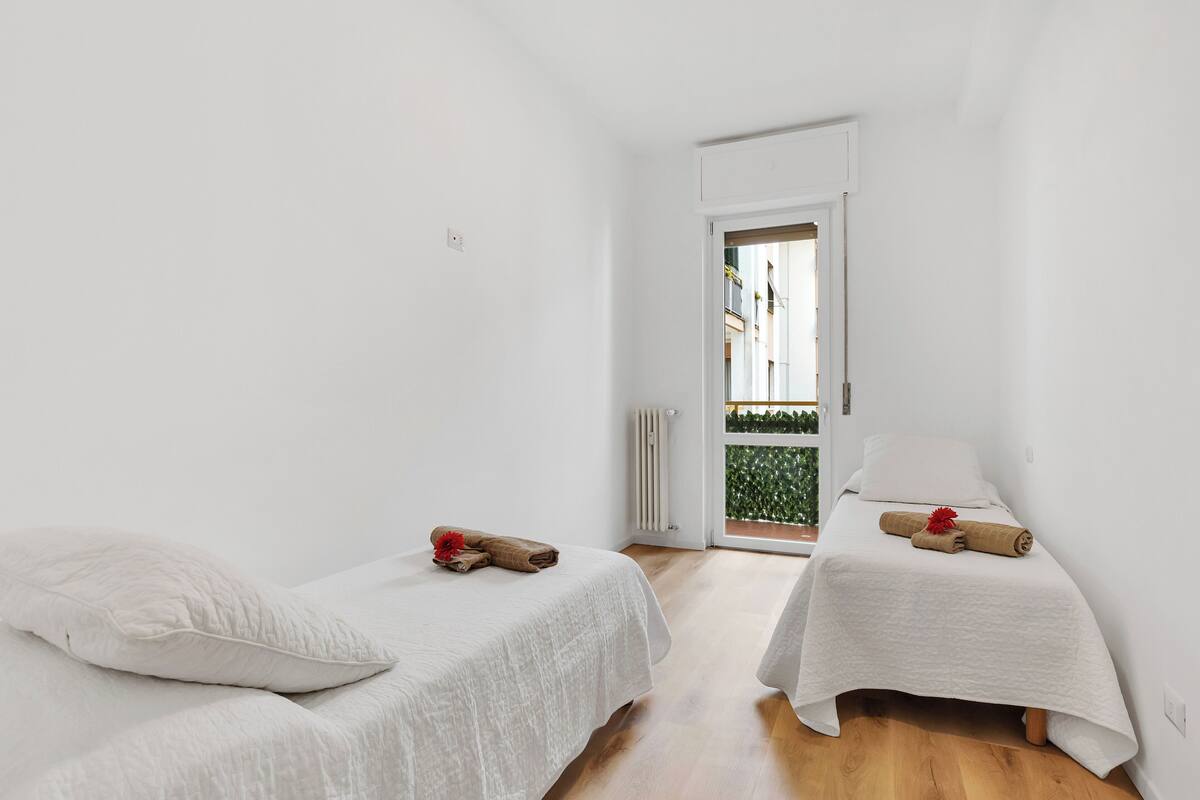 Quarto pequeno e iluminado com duas camas de solteiro, roupa de cama branca e vista para uma varanda com vegetação através de uma porta.