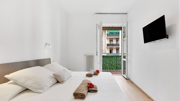 Une chambre d’hôtel moderne avec un lit soigneusement fait, un téléviseur à écran plat fixé au mur et une vue sur un immeuble doté de balcons, visible à travers une porte ouverte.