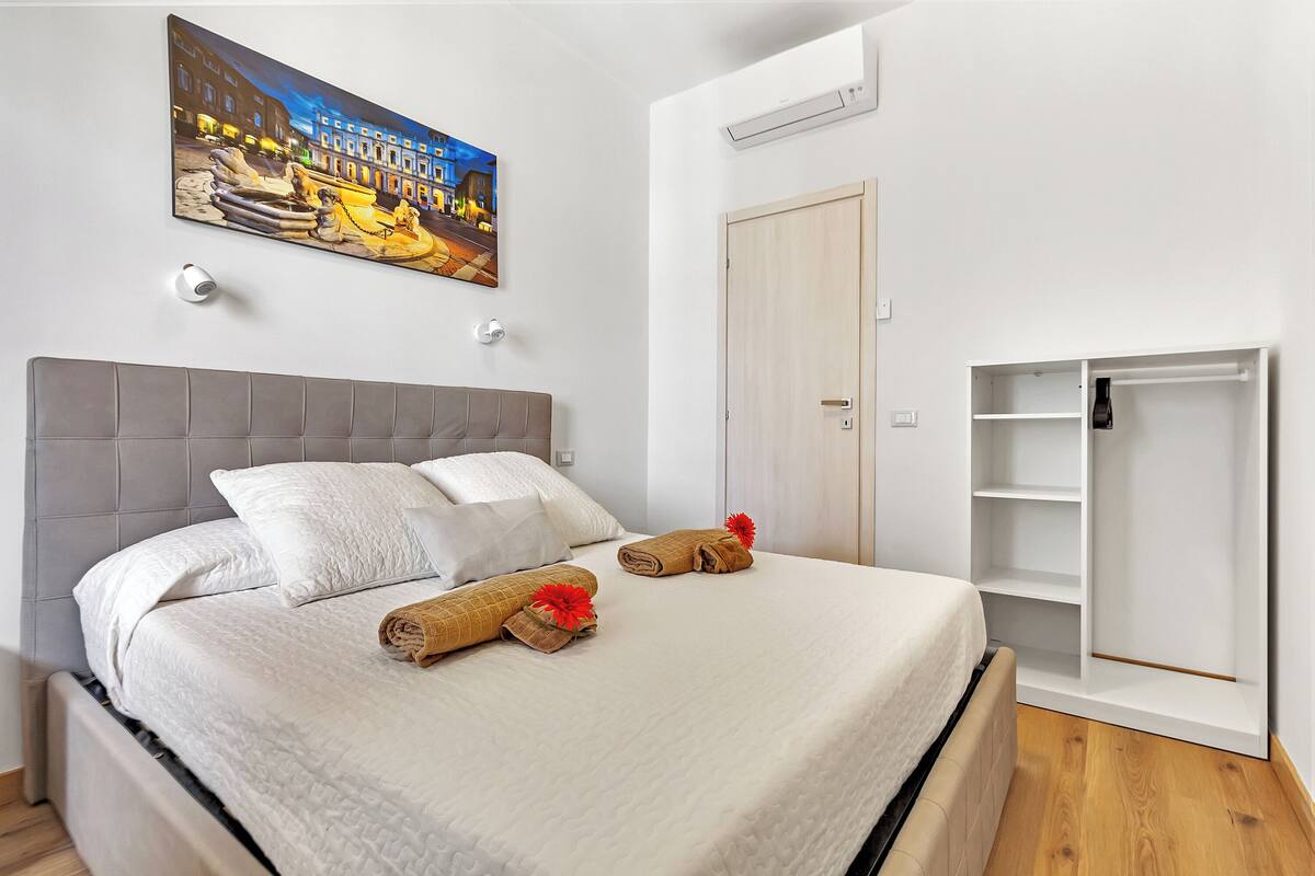 Cama bem arrumada com lençóis brancos e duas toalhas enroladas com flores vermelhas, um quadro com vista urbana acima, e um guarda-roupa com prateleiras abertas.