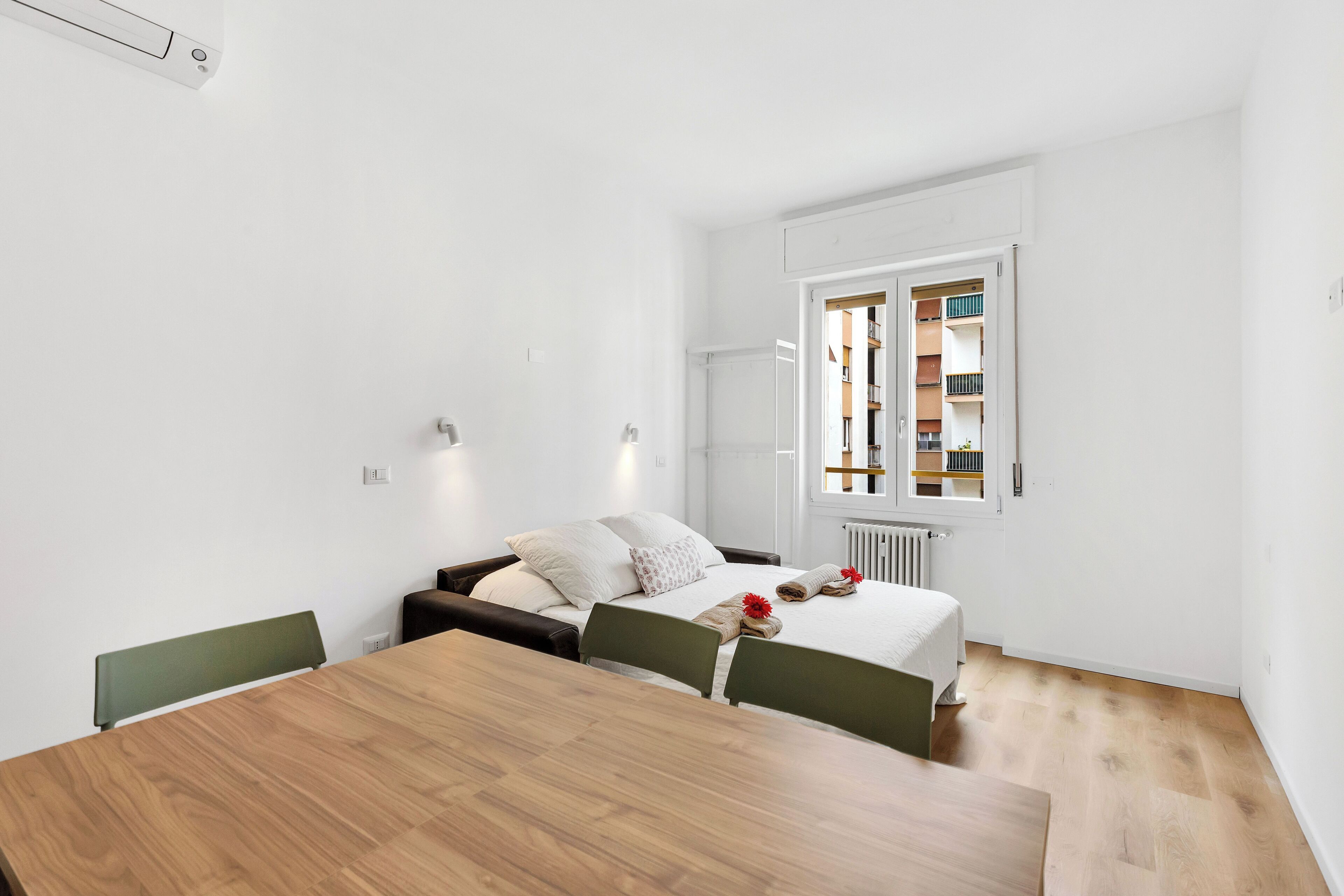 Una habitación de hotel moderna con una mesa de comedor de madera, una cama con ropa blanca y vistas a un edificio a través de una ventana.