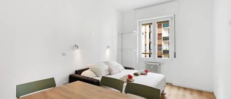 Una habitación de hotel moderna con una mesa de comedor de madera, una cama con ropa blanca y vistas a un edificio a través de una ventana.