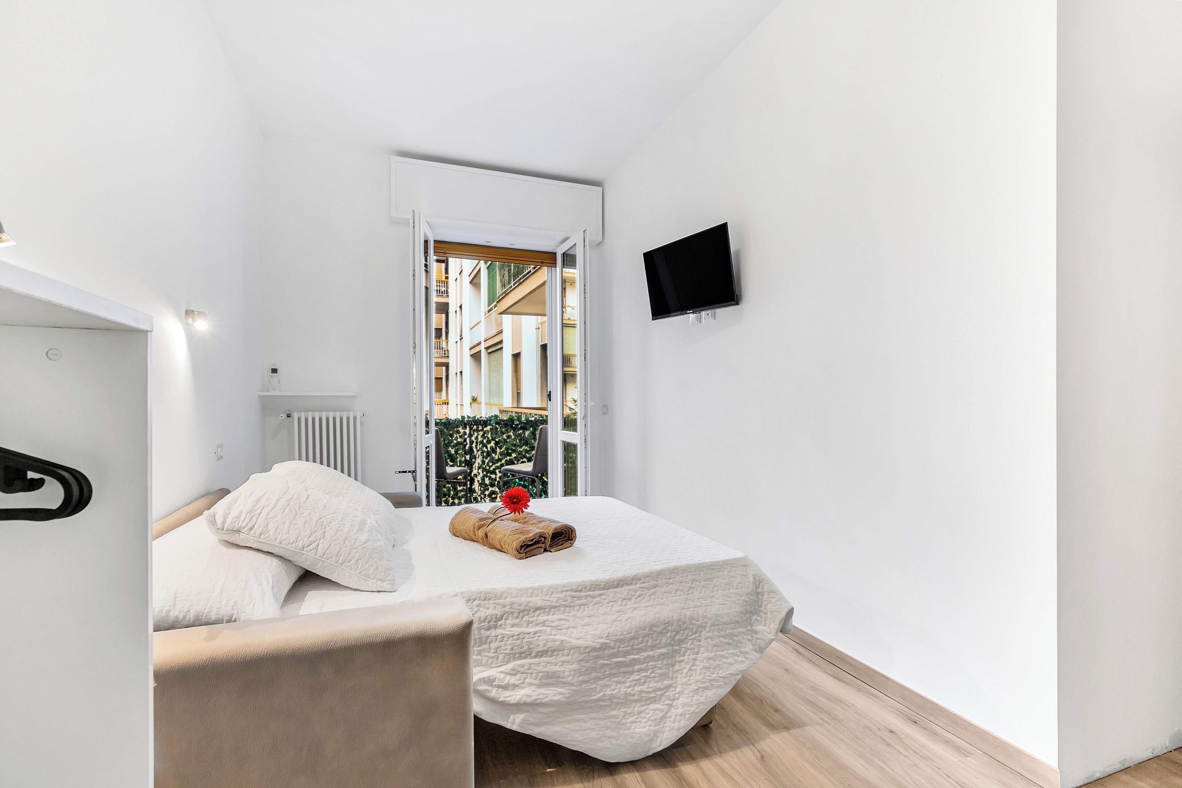 Un dormitorio con una cama, un televisor montado en la pared y una ventana con vista a edificios.