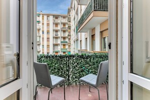 Een open balkon met twee stoelen, uitzicht op een meerhoog gebouw en een groene haag.