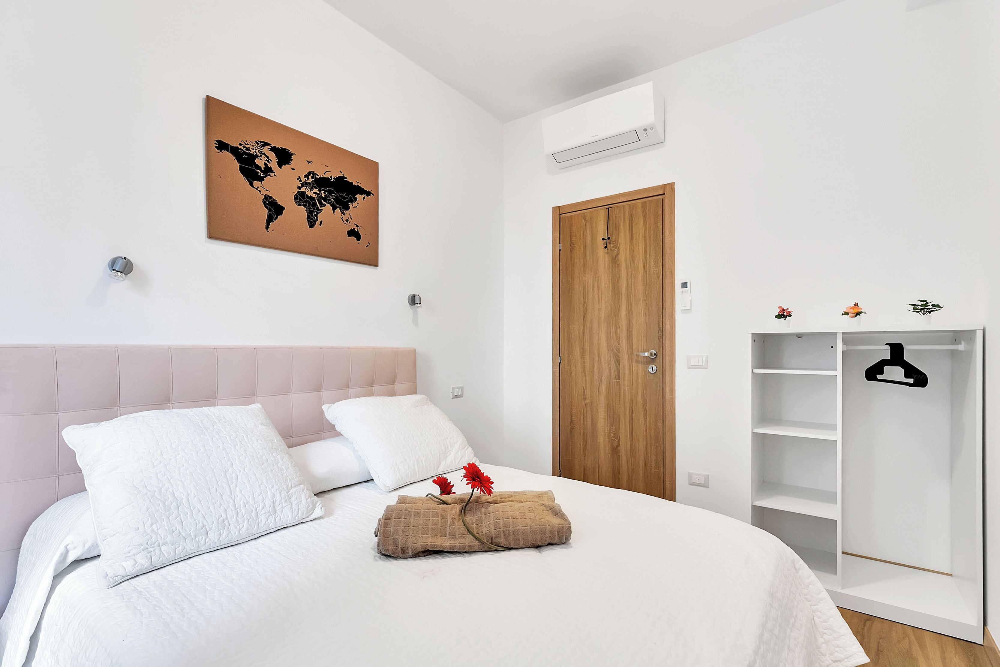 Ein ordentlich bezogenes Bett mit weißer Bettwäsche, ein Holz-Kopfteil, ein Weltkarten-Bild an der Wand und ein weißer Kleiderschrank mit Kleiderbügel.