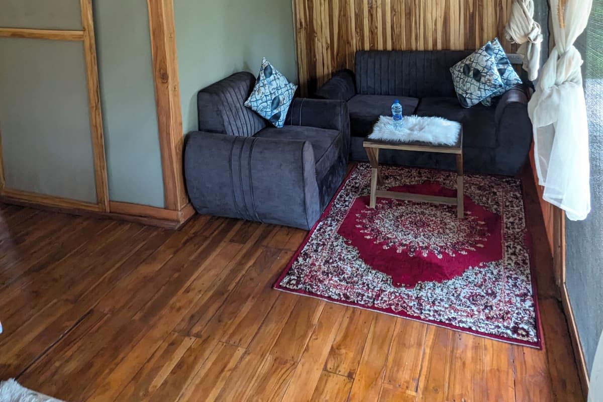 Een gezellige kamer met een houten vloer, een vloerkleed met een rood-wit patroon, een donkergrijze bank en een klein houten tafeltje met een waterfles.