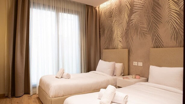 Ein Hotelzimmer mit zwei Betten, einem großen Fenster mit durchsichtigen Vorhängen, einem Kopfstück mit Palmenblattmuster und einem Nachttisch mit einem Buch und einer Kerze.
