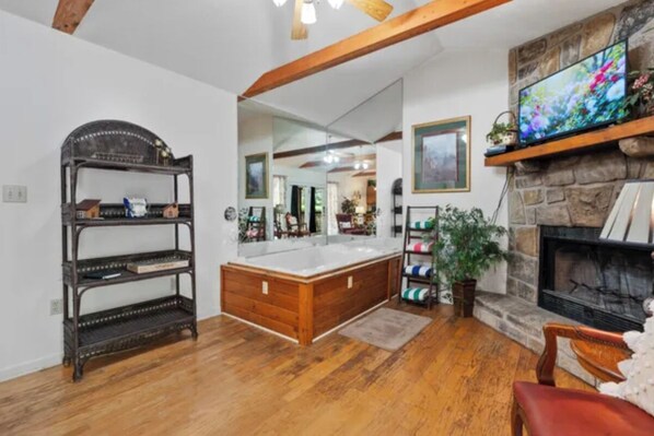 A room with a wooden floor, a stone fireplace, a large mirror, and a television on a stone shelf.